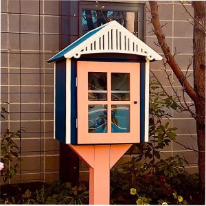 An outdoor wooden box with a hinged door, filled with books. The box is mounted on a post in a grassy area with trees in the background.
