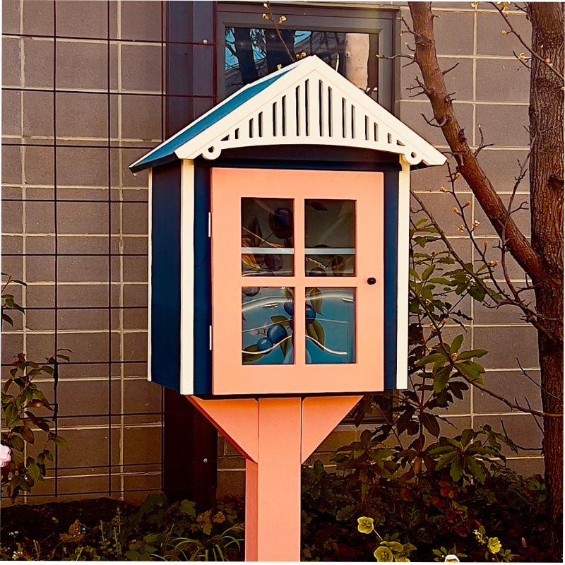 An outdoor wooden box with a hinged door, filled with books. The box is mounted on a post in a grassy area with trees in the background.